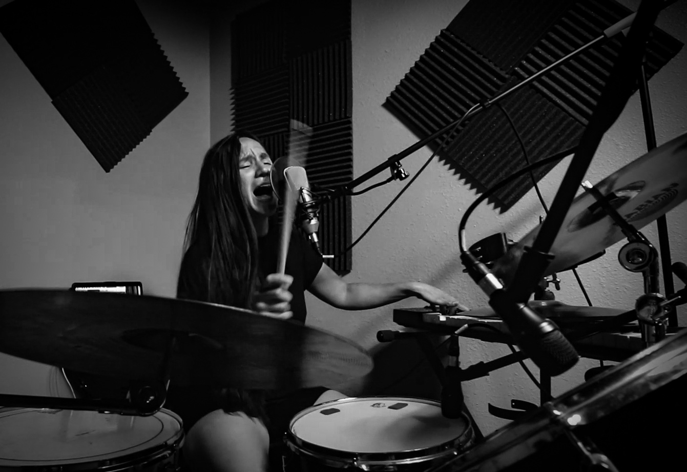 Madame singing into a microphone while playing drums and reaching across to a keyboard